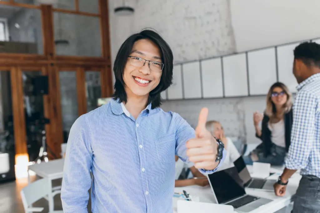 asian laughing boy posing with thumb up beginning workday chinese office worker blue shirt glasses smiling with laptop (2)