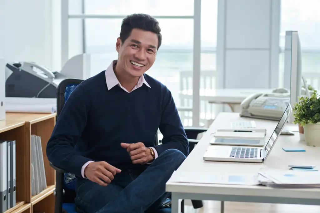 smiling asian man sitting desk front laptop office looking camera (1)