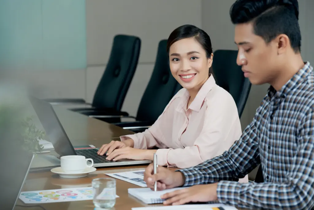 smiling asian woman sitting meting table with laptop amd male colleague writing notebook (1)