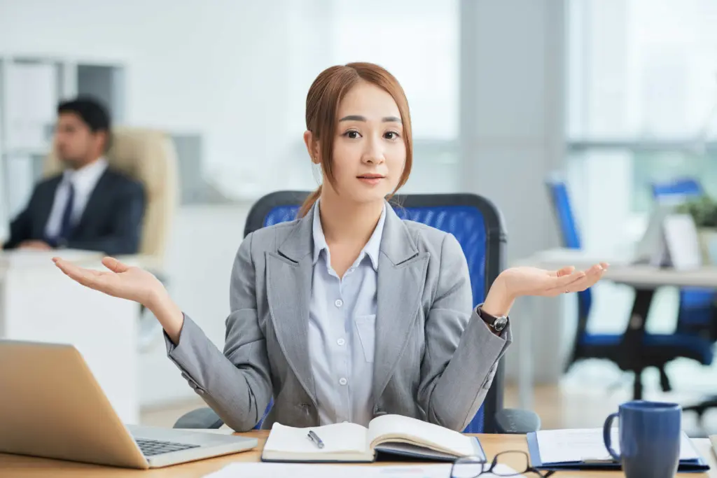 asian woman sitting desk office looking camera with helpless hand gesture (1)